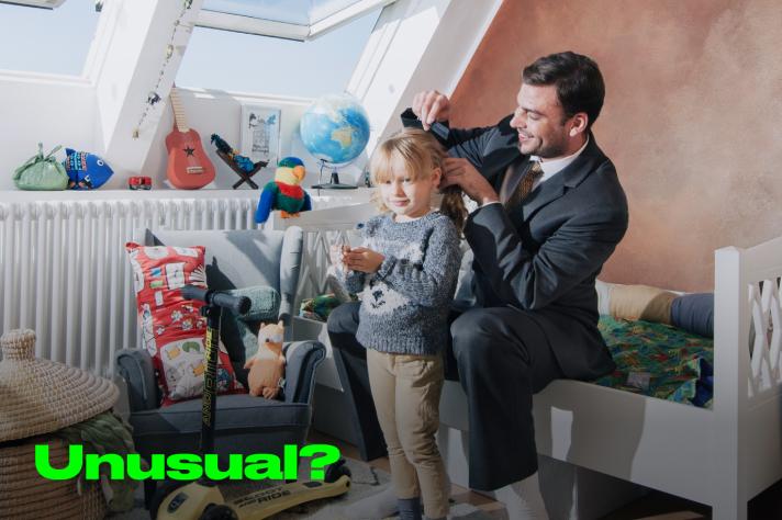 A working father dressed in a suit combing and styling the hair of his daughter with caption 'Unusual?'