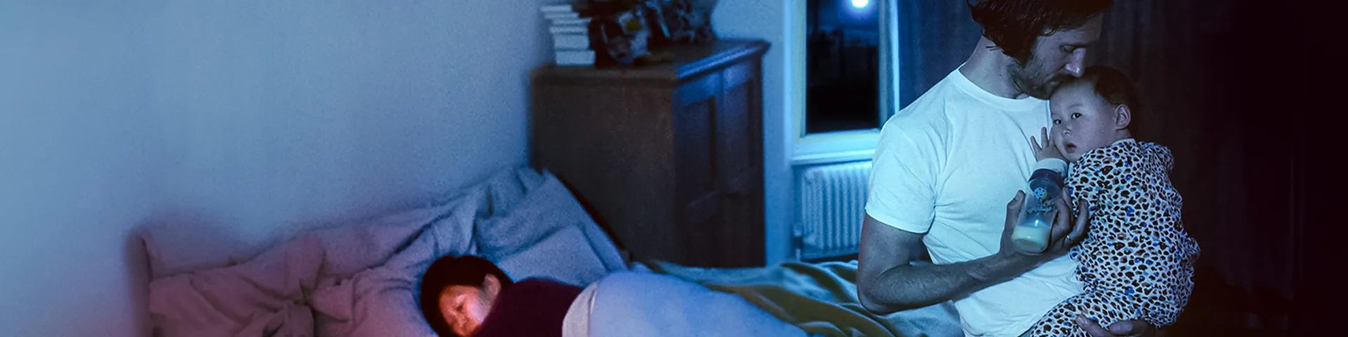 A father feeding a baby while mother is sleeping