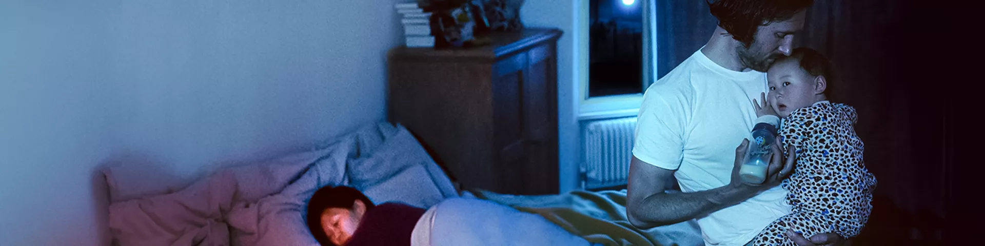 A father feeding a baby while mother is sleeping